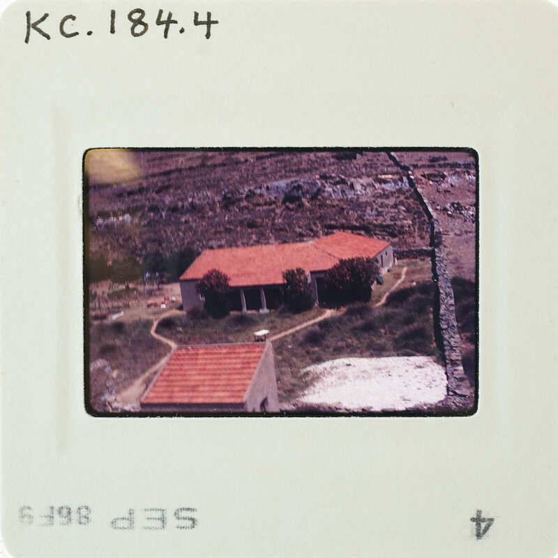 A faded photograph of a house with a red roof and a stone wall, taken from an aerial perspective.