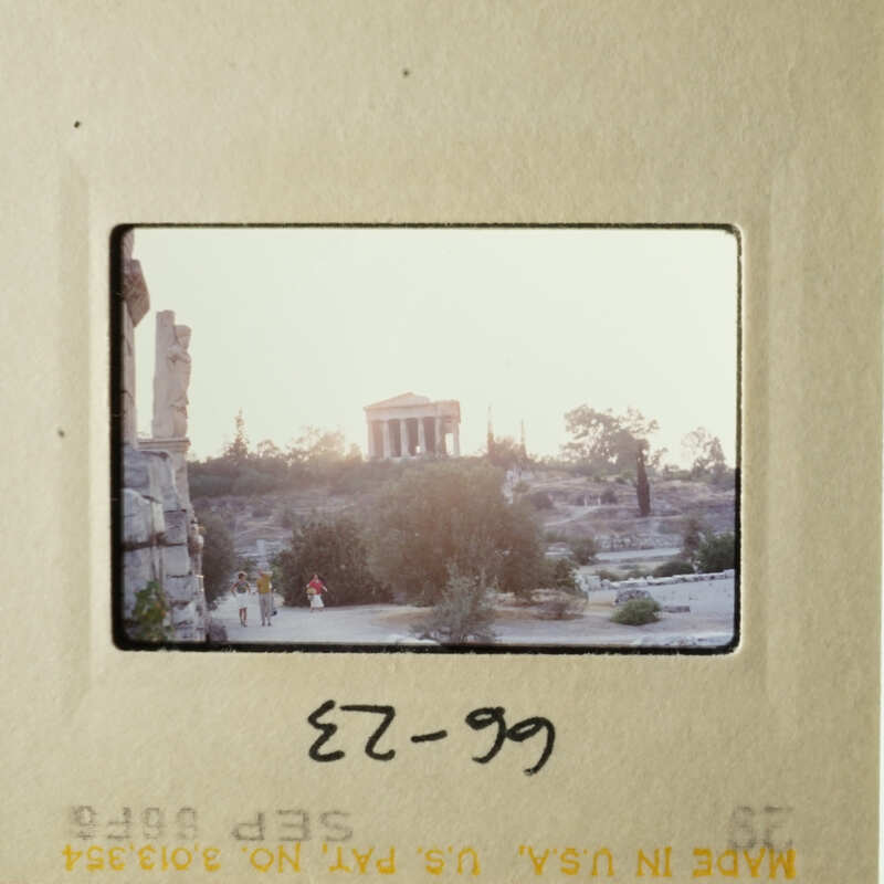 A photograph of an ancient Greek temple at sunset with people walking around it, as seen through a projector.