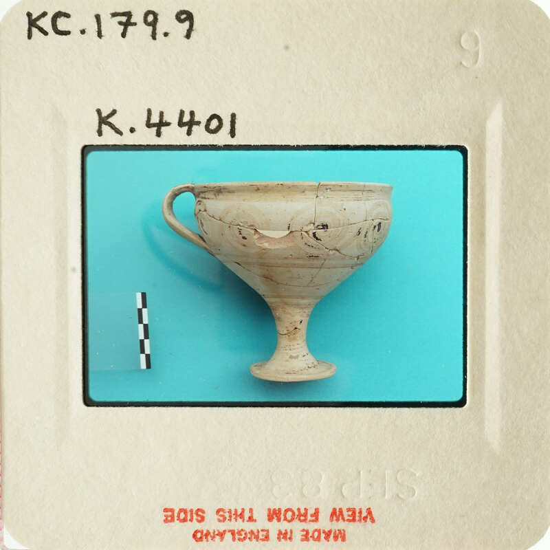 Ancient Greek cup with handle and face design, photographed on blue background.