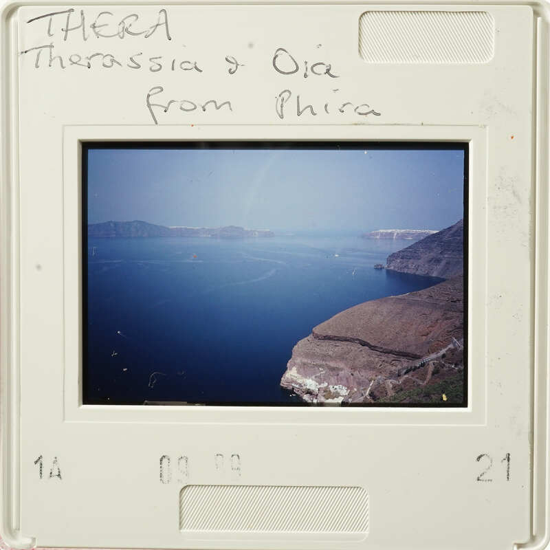 A photograph depicting a coastal landscape with a body of water and rugged terrain, possibly from Thera/Santorini, Greece.