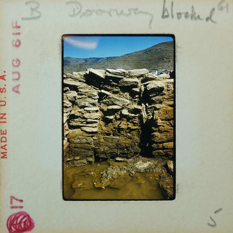 Detail of stone wall with doorway filled with similar stones, with mountains and sky in the background.