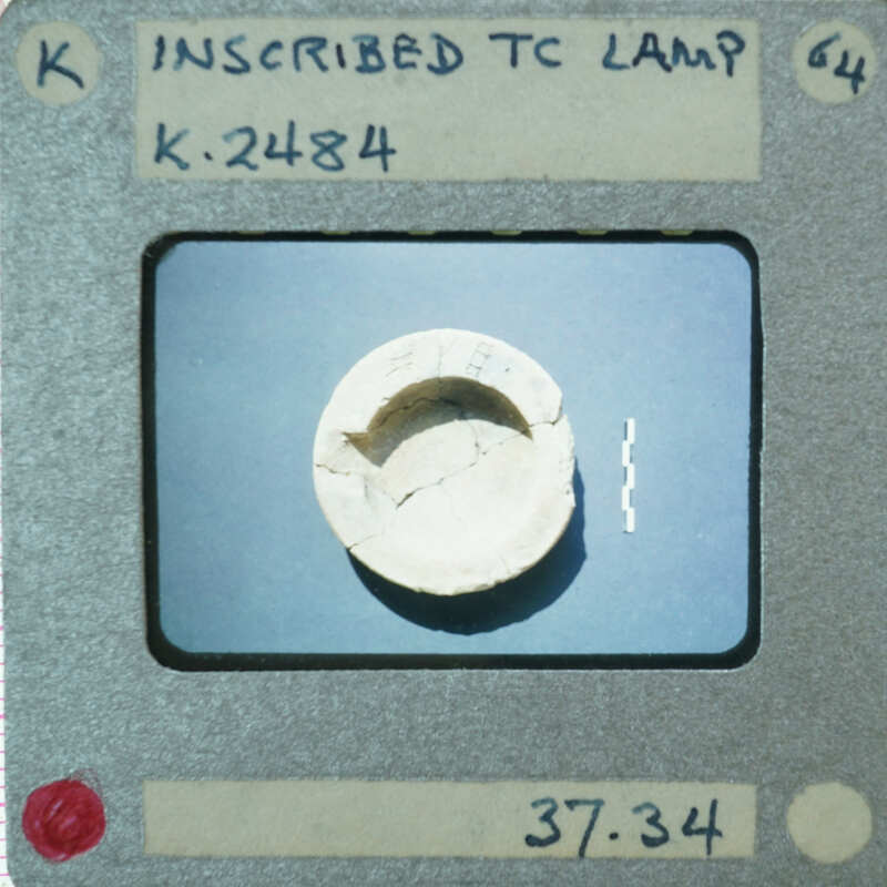 A small, circular object with a hole in the center, photographed against a gray background and labeled as "Inscribed TEC Lamp K-23481" on an old Polaroid film strip.