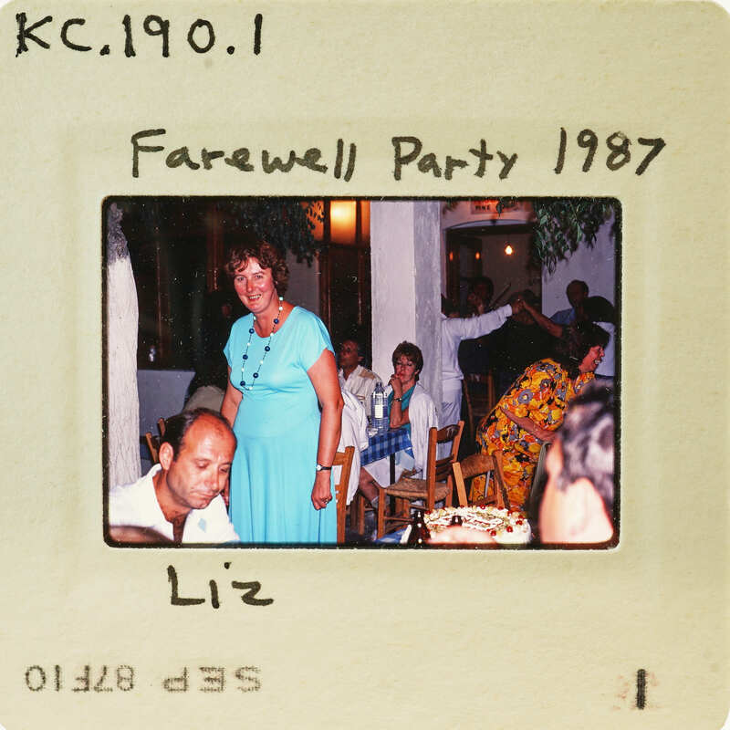 A woman standing at a party with people sitting around a table, as seen through an old photograph. Catalog number KC.190.1