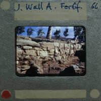Old photograph of a stone wall with trees and sky in the background, on a film strip with handwritten details.