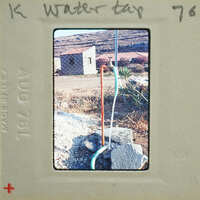 A photograph of a water tap with pipes and a stone base, set against a backdrop of a building and vegetation.