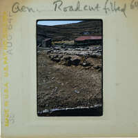 An old photograph of a dirt road with buildings and mountains in the background, taken on August 1967.