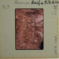 A close-up image of a bronze knife with an intricate handle, displayed on a card.