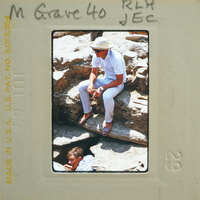 Man sitting on rocks with another man standing below him, wearing a hat and sunglasses.