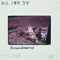 A photograph of a scenic coastal town with a prominent white building and houses overlooking the sea, labeled "Koundouros".