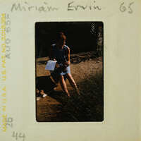 A vintage photo of a woman sitting on the ground with plants around her, wearing a sleeveless dress and holding papers or a book.