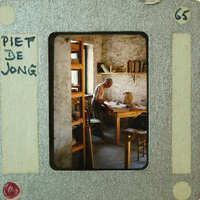 Older man sitting at a desk with books and papers, framed as an old photograph.