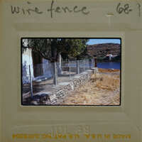 A photograph of a wire fence and stone wall on a hillside with trees and mountains in the background.