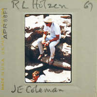 Man sitting on rock with another person behind him, wearing a hat and light-colored clothing.