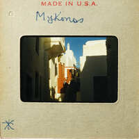 Old photograph of a narrow street with buildings and blue sky, labeled "Mykonos" on a cardboard mount.