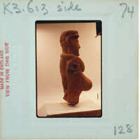 A photograph of an ancient clay figurine with a prominent belly, displayed on a stand.