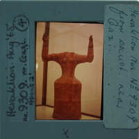 A photograph of a clay figurine with two arms and a headless body, displayed behind glass.