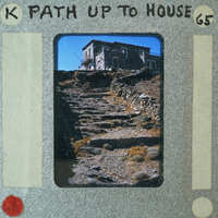 A photograph of a stone house with a path leading up to it, set against a mountainous landscape.