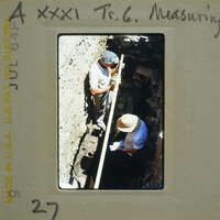 Two people working on a archaeological site with excavated earth and ruins visible.