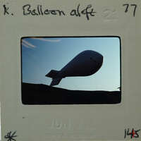 A black and white photograph of a large, round balloon with a tail flying against the sky.