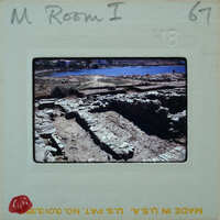 An old, partially excavated archaeological site with stone ruins and a body of water in the background.