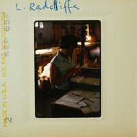A vintage photograph of a woman sitting at a desk with papers and a pen, wearing a light-colored blouse.