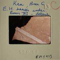 The photograph shows an ancient artifact, possibly a shard of pottery with visible cracks and details, set against a dark background.