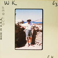 A man standing with his arms crossed, wearing a hat and shorts, near an old stone wall by the ocean.