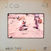 A man sitting on the ground, looking at something small in his hands while another person stands nearby.