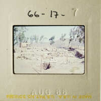 This photo shows an old, faded photograph of a barren landscape with a dirt road and some small structures, possibly ruins, under a clear sky.