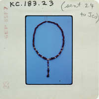 The image shows a bead necklace with a pendant against a blue background, with a white label containing handwritten notes and a barcode.