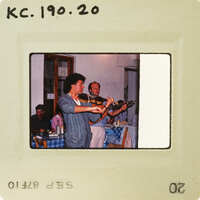 A vintage photo of a man playing the violin in front of a restaurant with checkered tablecloths and patrons seated at tables. Catalog number KC.190.20