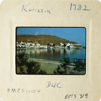 A photograph of an island village with houses and boats along the shore, taken from a distance.