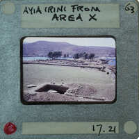 This is a vintage photo of an archaeological site with ruins and mountains in the background, presented on a film strip card labeled "AYA IRINI FROM AREA X".
