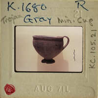 This is an image of a small, dark-colored cup with handles, possibly made of clay, displayed on a light surface.