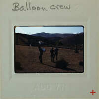 Three people standing on a field with mountains in the background, holding onto ropes attached to a hot air balloon.