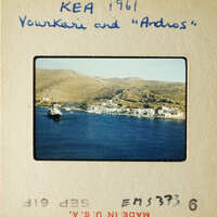 A vintage photograph depicting a coastal town with boats on the water, set against a backdrop of mountains and clear skies.