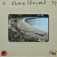 A photograph of a littered beach with trash scattered on the sand and water nearby.