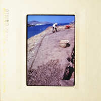 A person working on a dirt path near the sea, with a basket and a shovel nearby.