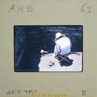 Person sitting on a chair outdoors, wearing a hat and measuring a pot.