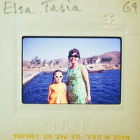 Two women posing together on a pier with water and mountains in the background.