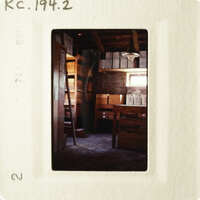 A photograph depicts an empty room with wooden walls and shelves, featuring boxes and a ladder.