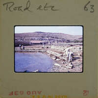 A vintage photo of a man working on a stone wall by a river, with mountains in the background.