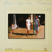 Two women standing together, smiling at the camera with a young child and an adult male behind them.