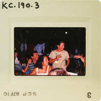 A vintage photograph of a group of people gathered at an event, with some individuals standing and others seated around tables. Catalog number KC.190.3