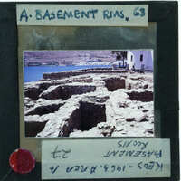 A photograph of a historical stone structure, possibly ruins, with the sea and mountains in the background.