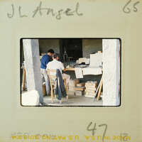 Old photograph of two men working at a table with various items, possibly tools and materials, set against the backdrop of an unfinished building or workshop.