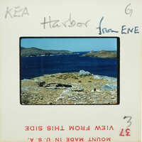 The image is a photograph of a scenic harbor with blue water, rocky shoreline, and distant islands under a clear sky.