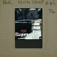 An old photo of a stone well with a sign on top, surrounded by ruins and greenery.