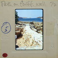 Slideof a stone wall leading to the sea with ruins and a clear sky.