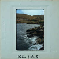 A vintage photo of a rocky coastline with crashing waves and distant hills.